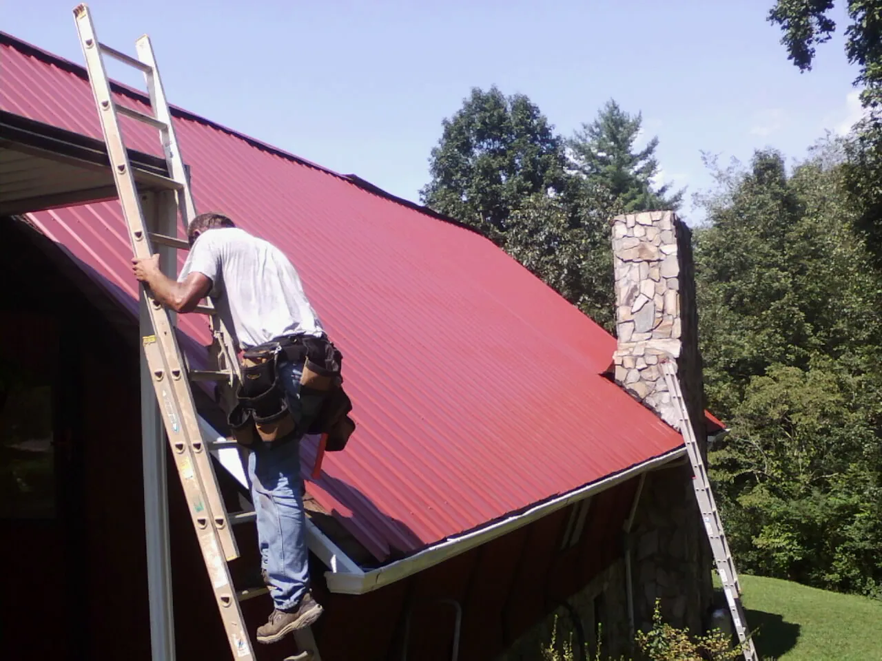 Roofer on a ladder beside a red metal roof and stone chimney — Soffit & Fascia Repair by Roofing Brothers in Bantry
